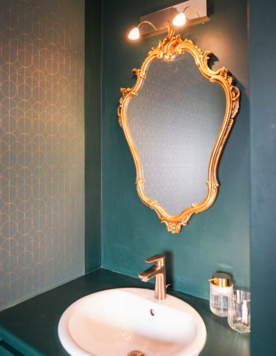 Salle de bain 30's avec miroir doré et robinetterie dorée. Décoration dans les tons vert et doré avec tapisserie à motifs géométriques
