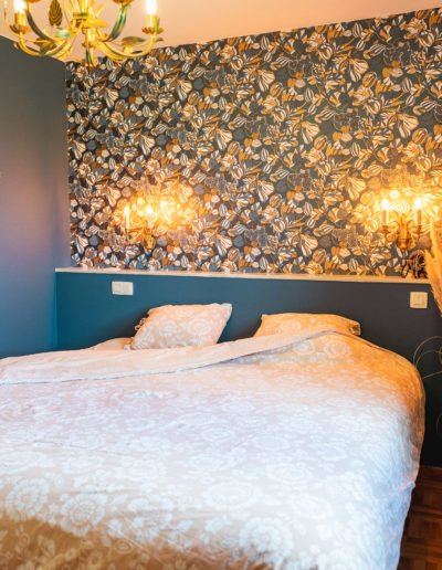 Chambre bleue avec lit 2 personnes et une tête de lit décorée d'une tapisserie à fleurs blanches et dorées du style art nouveau. Lustre type chandelier doré et appliques murales florales. Au mur, 3 miroirs dorés anciens